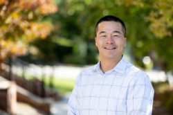A smiling man with short black hair stands outside in a white, light-blue checkered shirt. Fall trees and a sidewalk create a warm and vibrant background.