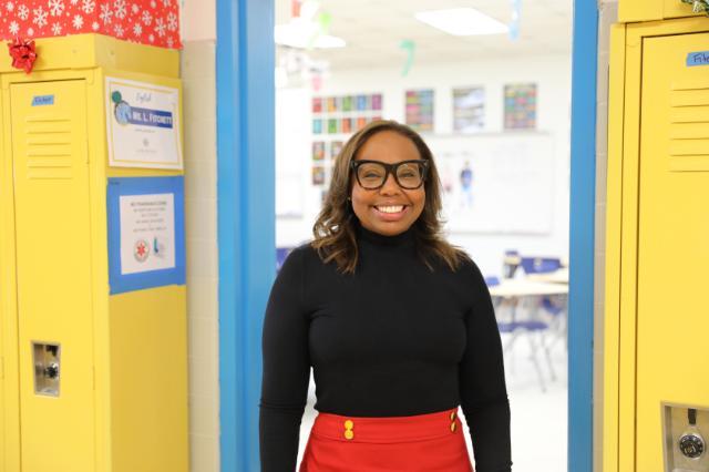 Lalisha Fitchett stands smiling in the doorway of her brightly colored classroom, framed by yellow lockers and a blue door. She is wearing glasses, a black long-sleeve top, and a red skirt with decorative yellow buttons. A sign on the locker identifies her as an English teacher. The classroom behind her is decorated with student work and colorful posters.