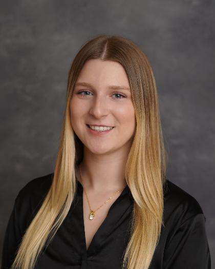 A young woman with long blonde hair is smiling in a professional headshot. She is wearing a black blouse and a gold necklace with a pendant. The background is a neutral gray, giving the image a formal and polished appearance. Her expression is warm and confident.