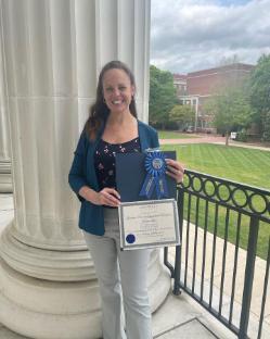 Kathryn Ticknor stands against a column and iron fence holding her first place blue ribbon.